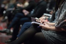 Multiple people sitting with their legs crossed taking notes at a conference.
